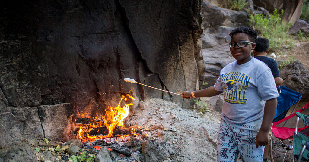 A young camper roasts a marshmallow over an open-air fire pit.